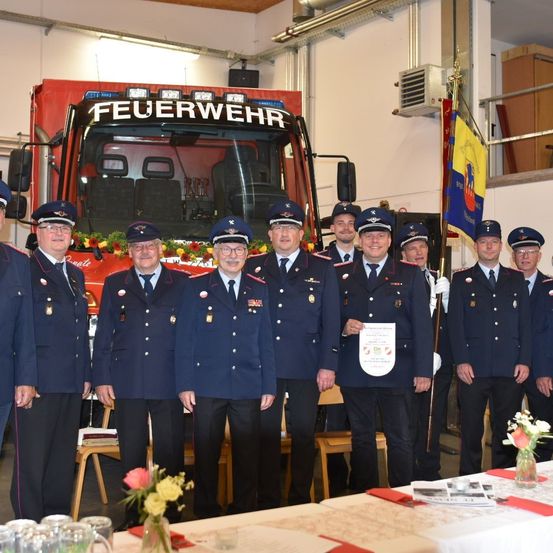 Eine Gruppe von Feuerwehrleuten in Uniform steht vor einem Feuerwehrauto mit Blumen darauf. Sie sind für eine offizielle Veranstaltung versammelt.