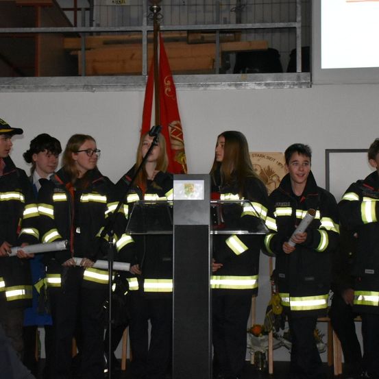 Eine Gruppe junger Menschen in Feuerwehruniformen steht vor einem Podium mit Mikrofon und hält gerollte Papiere. Ein Fahnenhintergrund ist sichtbar.