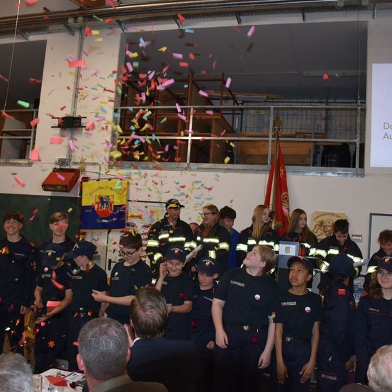 Eine Gruppe junger Menschen in Uniformen steht in einem Raum, in dem bunte Konfetti fallen. Sie schauen nach oben zur Decke. Einige von ihnen tragen Mützen und Brillen. Eine Fahne ist rechts. Es sitzen Leute auf der Seite. Eine Tafel ist an der Wand.