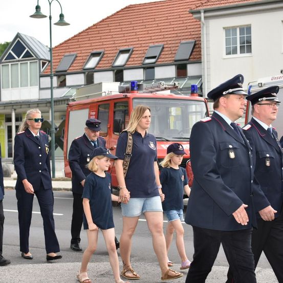 Eine Gruppe von Feuerwehrleuten in Uniform geht, begleitet von Kindern, über eine Straße. Ein roter Feuerwehrwagen ist hinter ihnen geparkt.