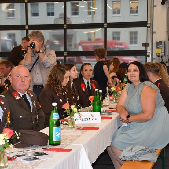 Eine Gruppe von Personen in Militäruniformen sitzt an einem langen Tisch, möglicherweise bei einem Bankett. Der Tisch ist mit Blumen, Flaschen und Namenskarten geschmückt. Ein Mann macht ein Foto mit einer Kamera.