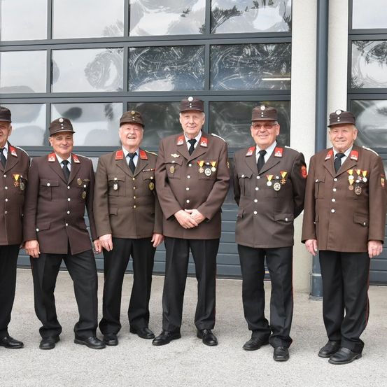 Fünf Männer in Militäruniformen stehen in einer Reihe und lächeln für die Kamera. Sie tragen Mützen, Medaillen und Krawatten. Der Hintergrund zeigt ein Gebäude mit Glaswänden.