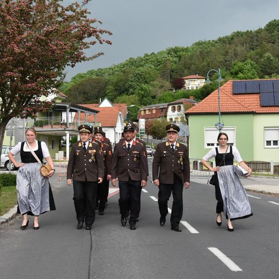 Eine Gruppe von Militäroffizieren und zwei Frauen in traditioneller Kleidung gehen eine Straße entlang, die von Häusern und Bäumen gesäumt ist. Die Häuser haben rote Dächer und einige verfügen über Solarpanels. Der Himmel ist bedeckt.