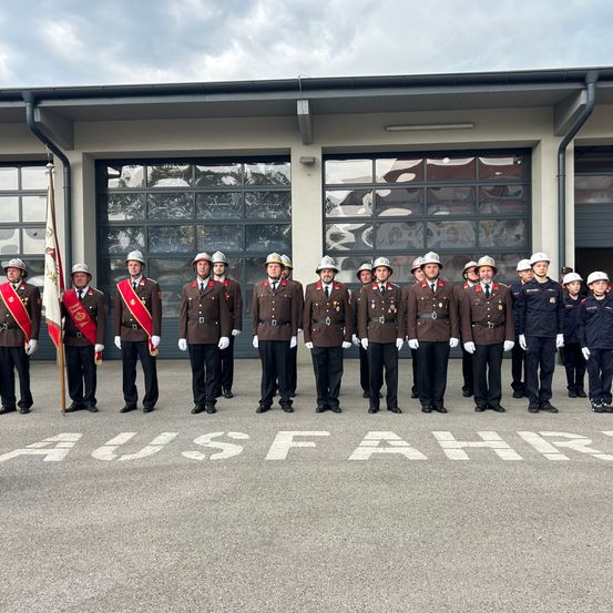 Eine Gruppe uniformierter Feuerwehrleute steht vor einer Feuerwache. Sie posieren für ein Foto, wobei das Wort 'AUSFAHR' auf dem Boden geschrieben steht.