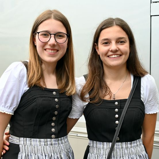 Zwei Frauen stehen zusammen und tragen beide traditionelle deutsche Kleidung. Die Frau auf der linken Seite hat eine Brille und eine Umhängetasche. Die Frau auf der rechten Seite trägt eine Halskette und hat ihre Hand auf der Schulter der anderen.
