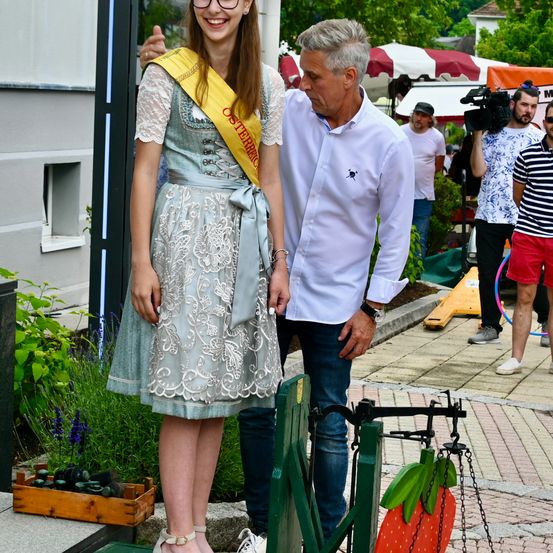 Eine Frau in einem traditionellen Kleid mit einer gelben Schärpe und ein Mann in einem weißen Hemd stehen draußen. Hinter ihnen filmen Leute mit Kameras, und einer hält eine Hula-Hoop.