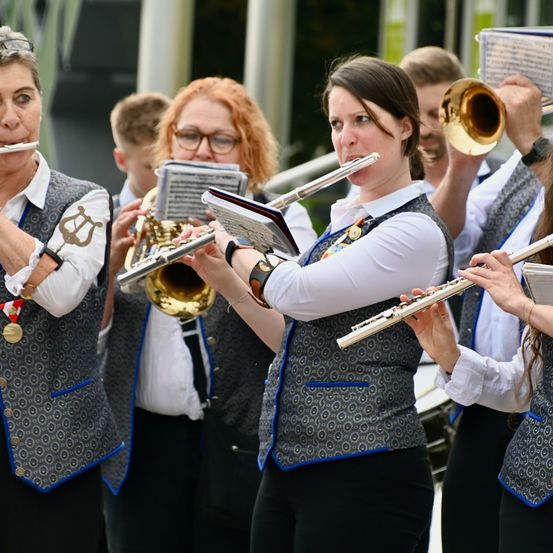 Eine Gruppe von Musikern, darunter Frauen, die Flöten und ein Mann, der eine Trompete spielt, führen gemeinsam in einer Außenumgebung auf.