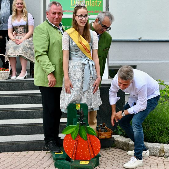 Eine Frau mit Krone und Schärpe steht auf einer Erdbeerwaage mit einem Mann und einer Frau hinter ihr. Ein anderer Mann beugt sich vor und inspiziert die Waage. Sie stehen auf einem Steinweg, mit Stufen und einem Gebäude im Hintergrund.