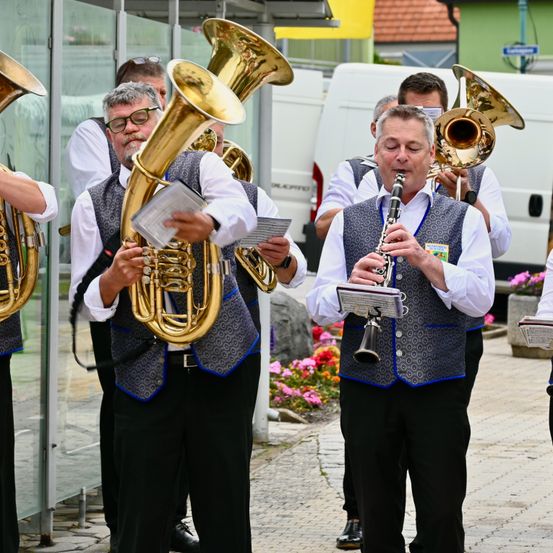 Eine Gruppe von Musikern spielt Blechblasinstrumente auf der Straße, darunter Tubas und eine Klarinette. Sie tragen Westen und stehen auf einem gepflasterten Bürgersteig.