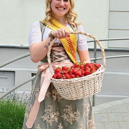 Eine Frau mit einer gelben Schleife mit dem Wort 'Erdbeere' hält einen Korb mit Erdbeeren und lächelt vor einem Gebäude.