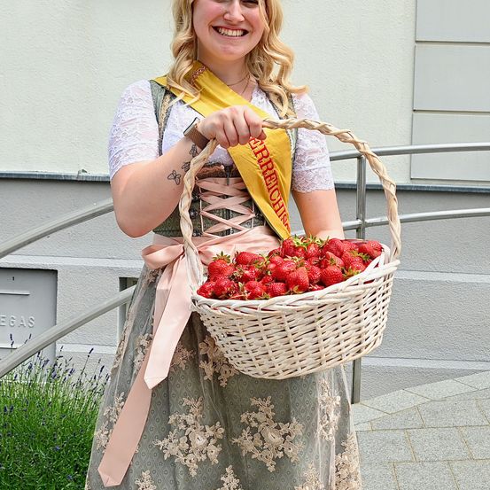 Eine Frau mit einer gelben Schleife mit dem Wort 'Erdbeere' hält einen Korb mit Erdbeeren und lächelt vor einem Gebäude.
