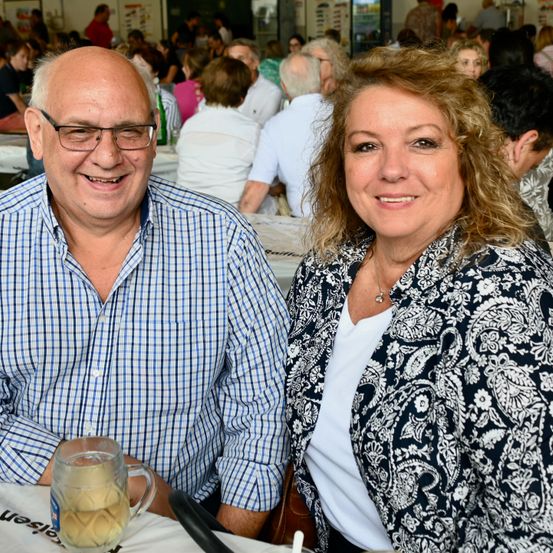 Ein älteres Paar sitzt an einem Tisch in einem überfüllten Bereich, lächelt und posiert für ein Foto. Der Mann hat ein Getränk in einem Glaskrug.