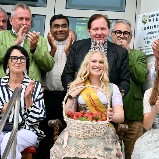 Eine Gruppe Erwachsener und eine Frau mit Krone, die einen Korb mit Erdbeeren hält, klatschen vor einem Gebäude.