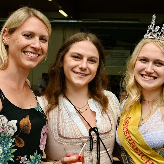 Drei Frauen stehen zusammen und lächeln für ein Foto. Die Frau auf der linken Seite trägt ein ärmelloses Blumenkleid, die Mitte hält ein Getränk und die Rechte trägt eine gelbe Schärpe.