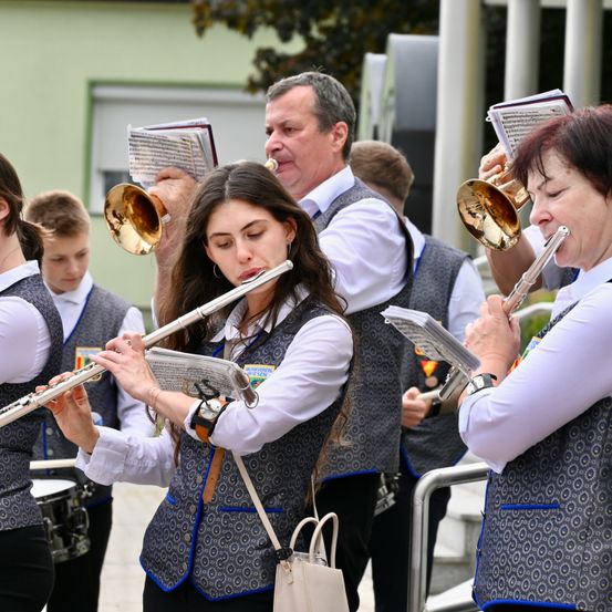 Eine Band probt draußen, wobei eine Frau die Flöte und ein Mann die Trompete spielt. Sie tragen beide gemusterte Westen und halten Notenblätter. Hinter ihnen spielt ein Junge die Trommel.