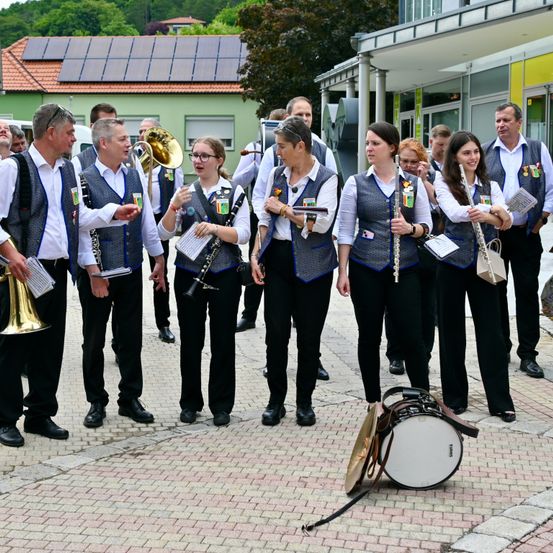Eine Blaskapelle versammelt sich vor einem Gebäude mit Solarmodulen. Die Bandmitglieder tragen Uniformen und halten Musikinstrumente. Ein Schlagzeug und Becken liegen auf dem Boden.