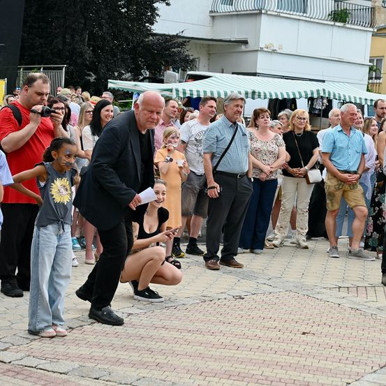 Eine Gruppe von Menschen steht um einen Mann herum, der ein Papier in der Hand hält. Er scheint zu sprechen. Die Menge ist in Alter und Geschlecht gemischt, einige machen Fotos.