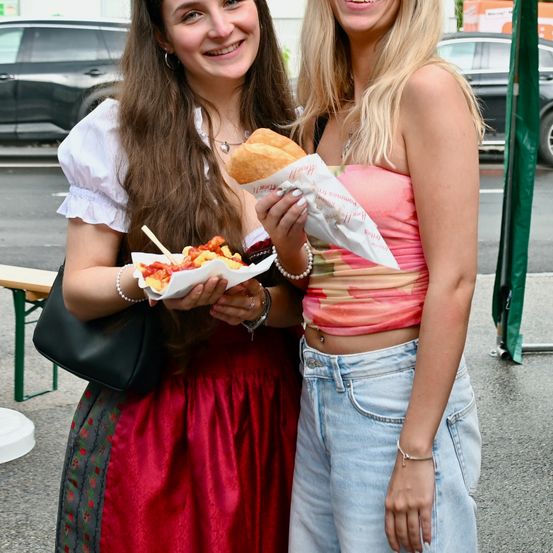 Zwei lächelnde Frauen, eine in einem roten Kleid und die andere in Jeans, halten einen Korb mit Essen und stehen draußen in der Nähe geparkter Autos.