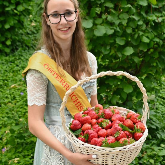 Eine Frau mit Brille trägt eine Schärpe und hält einen Korb voller Erdbeeren in einem Garten.