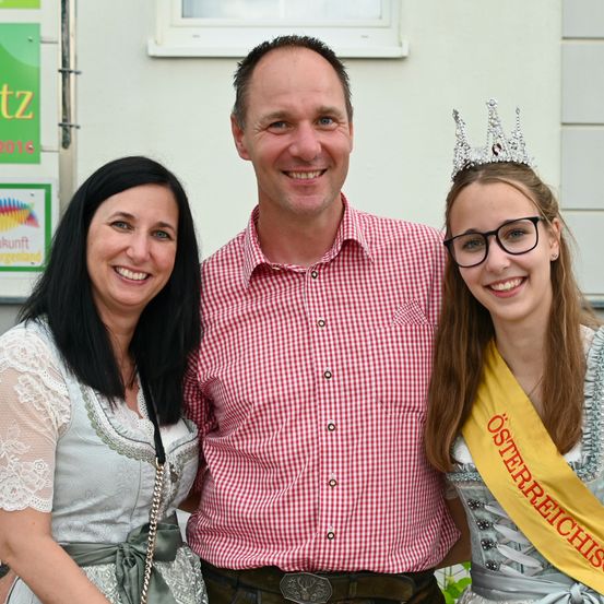 Ein Mann und zwei Frauen stehen nah beieinander und lächeln. Der Mann trägt ein rot-weiß kariertes Hemd. Die Frau auf der rechten Seite trägt eine gelbe Schärpe mit der Aufschrift 'Osterreichisch'. Die Frau auf der linken Seite trägt ein weißes Kleid mit einem grünen Band. Hinter ihnen steht ein weißes Gebäude.