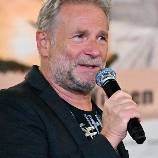 Ein mittelalte Mann mit Bart spricht in ein Mikrofon und trägt einen schwarzen Anzug. Er lächelt und scheint ein Publikum anzusprechen. Der Hintergrund ist unscharf.