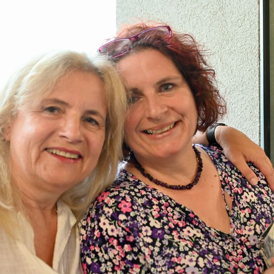 Zwei Frauen lächeln auf einem Foto, eine mit einer Brille auf dem Kopf, posieren für ein Bild mit einer weißen Wand im Hintergrund.