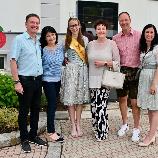 Eine Familie posiert draußen, wobei eine Frau eine Krone und einen Sash trägt, möglicherweise zur Feier eines Festivals.