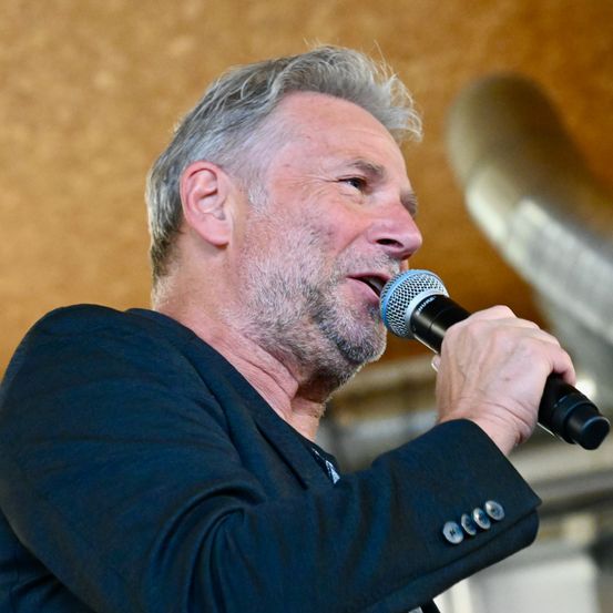 Ein Mann mit grauem Haar und Bart hält ein Mikrofon und spricht in dieses, während er nach vorne schaut. Er trägt einen dunklen Anzug.