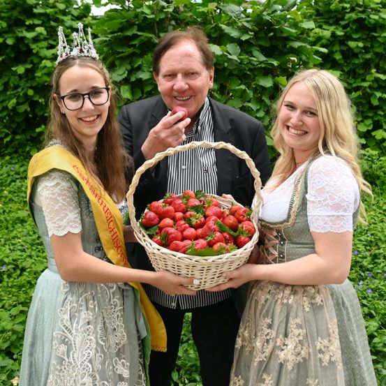 Drei Personen stehen in einem Garten mit einem Korb voller Erdbeeren. Ein Mann und zwei Frauen. Eine Frau trägt eine Krone und eine Schärpe, eine andere ein weißes Kleid mit einem grünen Oberteil.