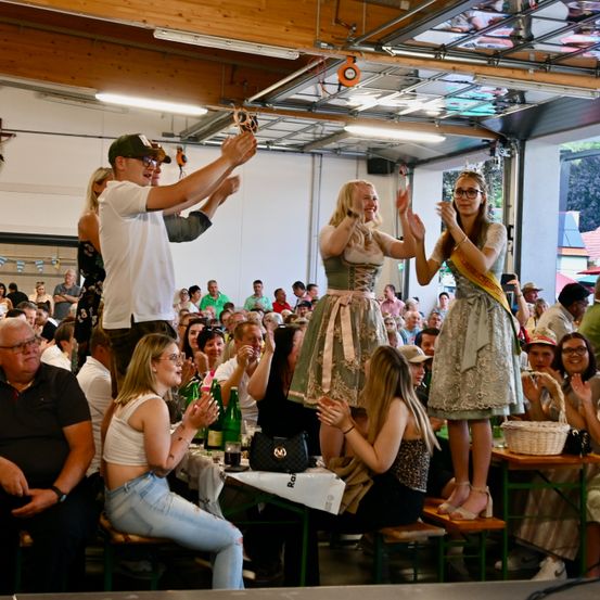 Eine Menschenmenge in einem Saal feiert ein Ereignis. Zwei Frauen stehen auf Stühlen, jubeln und heben ihre Hände. Hinter ihnen klatschen Menschen und halten Flaschen, vor ihnen steht ein Tisch.