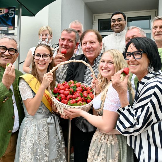 Eine Gruppe von Menschen versammelt sich draußen, lächelt und posiert mit Erdbeeren. Eine Frau hält einen Korb mit Erdbeeren, und eine andere Frau trägt eine Krone.