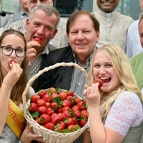 Zwei junge Frauen mit blonden Haaren genießen Erdbeeren in einem Korb, mit einem Mann und einem Priester hinter ihnen, alle lächeln bei einem sonnigen Event.