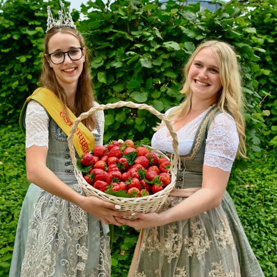 Zwei Frauen in traditioneller deutscher Kleidung, eine mit Krone, halten einen Korb mit Erdbeeren und lächeln fröhlich vor einem Hintergrund aus grünem Laub.