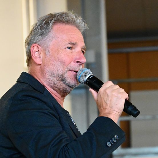 Ein Mann mit grauen Haaren und Bart spricht in ein Mikrofon. Er trägt einen schwarzen Anzug und hält das Mikrofon mit der rechten Hand.