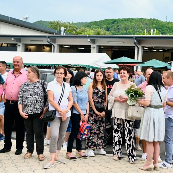Eine Gruppe von Menschen steht vor einem Gebäude, mit einem Berg im Hintergrund. Einige tragen eine Brille, und eine Frau hält einen Blumenstrauß. Vor dem Gebäude sind Autos geparkt.