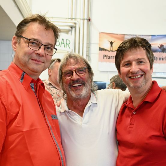 Drei Männer in orangefarbenen Hemden stehen gemeinsam und lächeln für ein Foto. Der Mann in der Mitte trägt eine Brille und ein weißes Hemd. Hinter ihnen zeigt ein Banner eine Person mit einem Sonnenuntergang im Hintergrund.