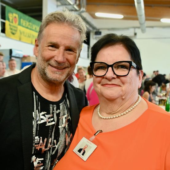 Ein älterer Mann und eine Frau lächeln für ein Foto bei einer Veranstaltung. Der Mann hat einen Bart und trägt ein schwarzes Shirt. Die Frau trägt eine Brille, eine Perlenkette und eine orange Bluse mit Ausweis. Im Hintergrund sitzen Menschen an Tischen.