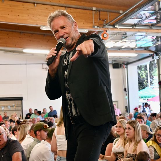 Ein Mann in einem schwarzen Anzug spricht in ein Mikrofon vor einer großen Menschenmenge. Das Publikum besteht aus verschiedenen Personen unterschiedlichen Alters, einige tragen Hüte und Brillen. Der Ort scheint eine Indoor-Veranstaltung zu sein.