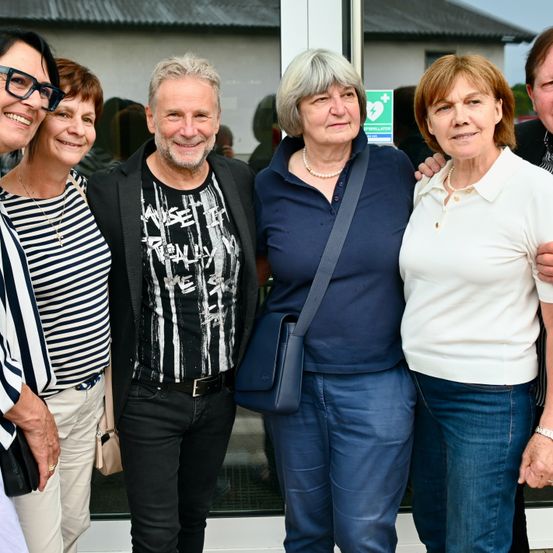 Fünf Personen, darunter zwei Frauen und drei Männer, stehen nebeneinander vor einer Glastür. Die Frau auf der rechten Seite trägt ein weißes Hemd und eine Halskette.