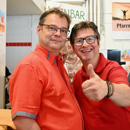 Zwei Männer in orangefarbenen Hemden posieren für ein Foto, einer gibt Daumen hoch. Sie stehen nebeneinander, lächeln, mit einem verschwommenen Hintergrund, der eine Wand und Menschen zeigt.