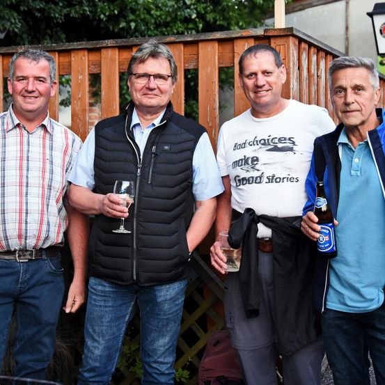 Vier Männer stehen zusammen in einem Garten. Einer hält ein Weinglas, ein anderer eine Bierflasche. Sie lächeln und scheinen für ein Foto zu posieren. Hinter ihnen ist ein Holzzaun zu sehen.