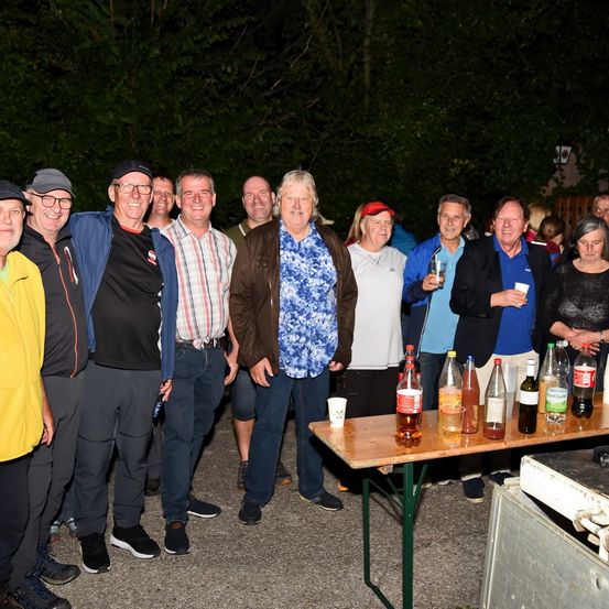 Eine Gruppe von Erwachsenen steht zusammen in einer Außenumgebung. Sie tragen Freizeitkleidung, und einige halten Getränke. Ein Tisch mit Flaschen und Gläsern steht vor ihnen. Bäume und eine Straßenlaterne sind im Hintergrund.