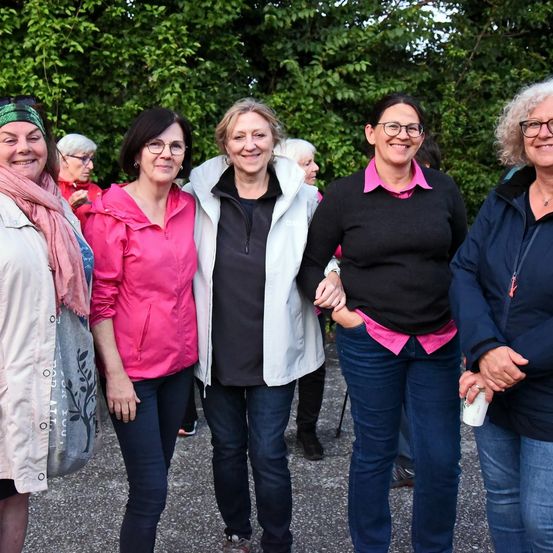 Fünf Frauen stehen eng beieinander in einem Park, lächeln und posieren für ein Foto. Die Frau auf der linken Seite trägt einen Schal. Bäume und Grünpflanzen sind hinter ihnen.