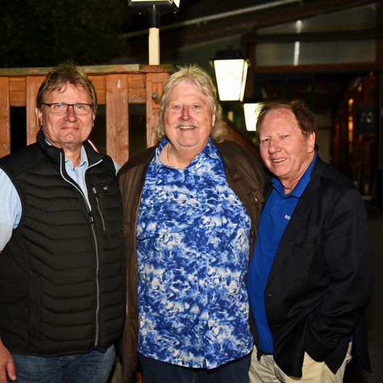 Drei Männer stehen nachts vor einem Holzzaun. Der Mann links trägt eine schwarze Weste, der mittlere ein blaues T-Shirt und der rechte ein blaues Hemd und eine schwarze Jacke.