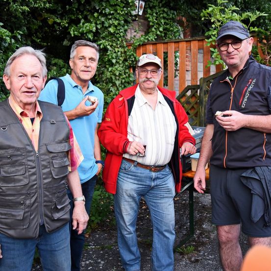 Vier Männer stehen draußen in der Nähe eines Holzzauns, lächelnd und Essen haltend. Ein Mann trägt eine graue Weste, ein anderer eine rote Jacke, ein dritter ein blaues Hemd und der letzte eine schwarze Jacke.