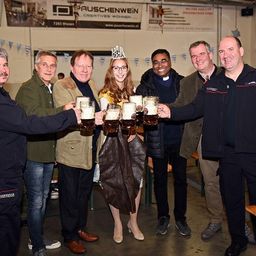 Eine Gruppe von Menschen in einem Saal, lächelnd und mit Tassen in der Hand, eine Frau mit Krone, mit Bannern im Hintergrund.