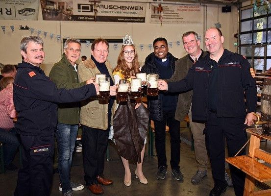 Eine Gruppe von Menschen in einem Saal, lächelnd und mit Tassen in der Hand, eine Frau mit Krone, mit Bannern im Hintergrund.