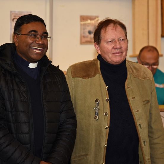 Zwei Männer, einer mit Brille und schwarzem Mantel, der andere in einer braunen Jacke mit einer Anstecknadel, stehen lächelnd für ein Foto. Hinter ihnen schaut ein Mann mit Brille zu.