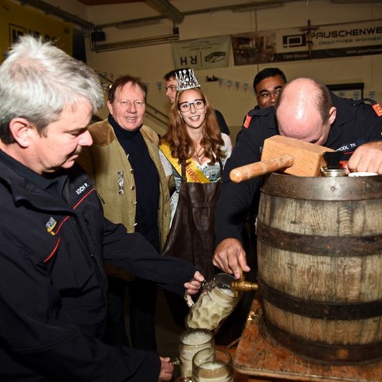 Eine Gruppe von Menschen steht um einen Holzfass herum, wo ein Mann einen Hammer benutzt, um einen Zapfhahn anzuklopfen. Ein anderer Mann zapft Bier aus dem Fass in Gläser ab.