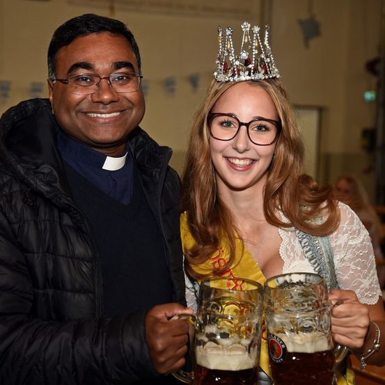 Ein lächelnder Priester und eine Frau mit Krone halten zwei Gläser Bier, wahrscheinlich auf einem Fest.